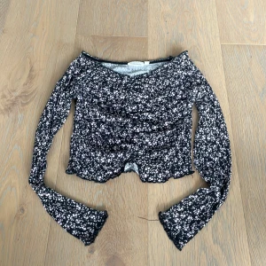 Blommig  topp från KappAhl - Supersöt croppad långärmad topp från KappAhl med svart botten och små vita och ljusrosa blommor. Toppen har v-ringning, ribbad struktur och vågig kant nertill och vid ärmslut. Perfekt för dig som gillar en  trendig stil.