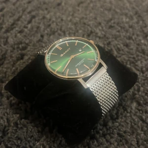 Grön GANT klocka med mesharmband - Stilren klocka från GANT med grön urtavla och silverfärgade detaljer. Klockan har ett klassiskt runt boett och ett elegant mesharmband i metall. Urtavlan har guldiga index och visare samt datumvisning. Perfekt accessoar för en snygg look.