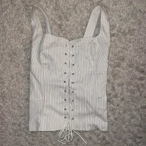 TRENDIG Vit randig korsett topp från Zara - Intressekoll! Supersnygg vit korsett-topp med tunna svarta ränder och breda axelband. Framsidan har cool snörningsdetalj hela vägen ner och baksidan har dragkedja för enkel påklädning. Perfekt för att lyfta vilken outfit som helst! Omsydd för o passa xxs-s! 