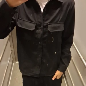 Svart overshirt från H&M - Säljer en svart overshirt från H&M med klassisk krage, två stora bröstfickor med lock och knäppning framtill. Skjortan är långärmad och har en clean, stilren look som passar till många outfits. Materialet är mjukt och känns bekvämt mot huden.