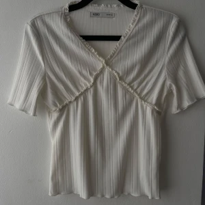 Vit ribbad blus med volang från Mixxo - Supersöt vit blus från Mixxo med ribbad struktur och korta ärmar. Toppen har v-ringning och fina volangdetaljer längs halsringningen och bysten. Perfekt för dig som gillar en romantisk och stilren look.