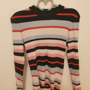Randig ribbad långärmad tröja - Säljer en ribbad långärmad tröja med färgglada ränder i svart, rött, rosa, ljusblått och beige. Tröjan har en tight passform och rund halsringning. 
