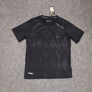 Svart tränings-tshirt från Under Armour - Svart kortärmad t-shirt från Under Armour med diskret geometriskt mönster och lila logga på bröstet. Tillverkad i ett lätt och snabbtorkande material som passar perfekt för träning. Klassisk rund hals och sportig vibe.