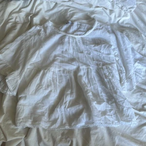 Vit blus med spets från Ellos - Luftig vit blus från Ellos i storlek 40. Blusen har rund halsringning med söt spetskant, vida ärmar och volangdetaljer. Tillverkad i ett lätt och skönt bomullstyg som passar perfekt för varmare dagar.