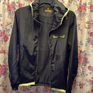 Svart vindjacka från Fendi med gula detaljer - Svart vindjacka från Fendi med huva och dragkedja. Jackan har coola gula detaljer vid huva, nederkant och på bröstet med Fendis ikoniska ögon. Tillverkad i glansig polyester som ger en sportig vibe. Perfekt för dig som vill sticka ut med designerstil.