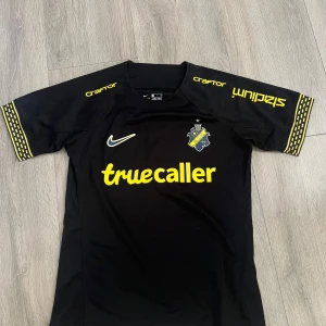 AIK matchtröja Milosevic - AIK:s officiella matchtröja från Nike i svart med gula detaljer och tryck. Nummer 5 och Milosevic på ryggen, klubbmärke på bröstet och sponsorer på ärmar och framsida. Tillverkad i Dri-Fit-material som andas, perfekt för fotbollsträning eller match.