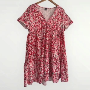 Blommig kortklänning - Supersöt kortklänning från SHEIN med röd- och rosa blommigt mönster på ljus botten. Klänningen har v-ringning, korta ärmar och en luftig passform som ger en chill vibe. Perfekt för varma dagar när du vill känna dig både söt och bekväm. Säljer för att den ej passar mig, i storlek M. Använd endast 1 gång.