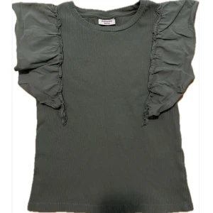 Olivgrön volangblus - Supersnygg olivgrön blus från Kappahl med ribbad struktur och volanger vid axlarna. Perfekt för dig som vill vara basic men ändå lite finare! Storlek 158/164 men passar XS. 