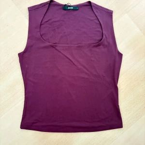 Vinröd ärmlös topp från Bik Bok - Vinröd ärmlös topp från Bik Bok i storlek L. Toppen har en rundad, djup urringning och är figurnära i modellen. Tillverkad i mjuk bomullsmix som känns skön mot huden. Perfekt att styla med jeans eller kjol för en clean look.