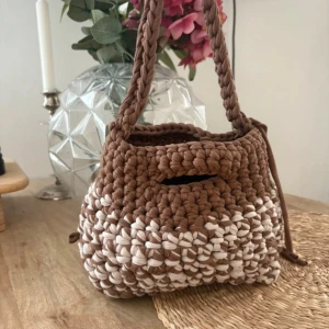 Handgjord virkad axelväska i brunt - Superfin handgjord axelväska i grovt virkad bomull med tvåfärgat mönster i brunt och beige. Väskan har en stadig flätad axelrem och dragsko i sidan för att stänga väskan. Perfekt för dig som gillar unika accessoarer med boho-vibe.