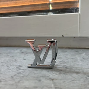 Louis Vuitton LV bältesspänne silver - Säljer ett ikoniskt LV bältesspänne från Louis Vuitton i blankt silverfärgat metall. Klassisk design med LV-loggan framtill och ingraverad text på baksidan. Perfekt för att ge din outfit en lyxig och stilren touch.