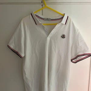 Vit Moncler pikétröja med ränder - Snygg vit pikétröja från Moncler med klassisk krage och knäppning framtill. Tröjan har röda och svarta ränder på krage och ärmslut samt Moncler-logga broderad på bröstet. Perfekt för en clean och sportig look.