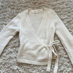 Beige omlottkofta från Gina Tricot - Supermjuk beige omlottkofta från Gina Tricot i ullblandning. Koftan har långa ärmar, ribbade kanter och knytband i sidan för en snygg passform. Köpt för 400 kr.