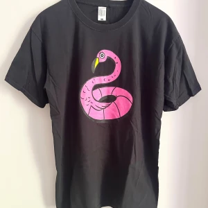 Lollapalooza Stockholm t-shirt svart L - Svart t-shirt från Fruit of the Loom i storlek L med färgglad flamingo-tryck på framsidan och Lollapalooza Stockholm 2022 lineup på baksidan. Klassisk passform och rund hals, tillverkad i mjuk bomull. Perfekt för dig som älskar festivaler och coola prints.