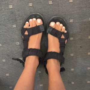 Svarta chunky sandaler med remmar - Säljer ett par svarta sandaler med breda, vadderade remmar och platt sula. De har öppen tå och justerbara spännen runt vristen. Perfekta för sommaren och riktigt bekväma att gå i. Snyggt chunky uttryck som passar till många outfits.