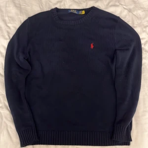 Mörkblå stickad tröja Polo Ralph Lauren - Mörkblå stickad tröja från Polo Ralph Lauren med klassisk röd broderad logga på bröstet. Tröjan har rund hals, ribbade muddar och är långärmad. Perfekt för dig som gillar stilren och tidlös design.