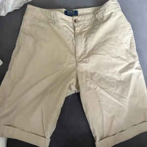 Beige shorts från Polo Ralph Lauren - Klassiska beige shorts från Polo Ralph Lauren med normal passform. De har fickor fram och bak, bälteshällor och stängs med knapp och dragkedja. Perfekta för varma dagar och enkel att matcha med det mesta. Det finns ett pyttelitet hål vid höger bakficka, men är knappt märkbart.
