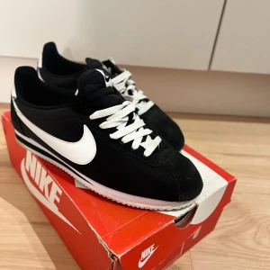 Nike Cortez svartvita sneakers - Klassiska Nike Cortez sneakers i svart och vitt med vit sula och tydlig Swoosh-logga på sidan. Skorna har snörning, rund tå och ovandel i syntet och mocka. Perfekta för dig som gillar retrostil och vill ha en ikonisk sneaker i garderoben.
