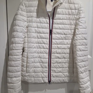 Vit quiltad pufferjacka Tommy Hilfer - Snygg vit pufferjacka med quiltad design och dragkedja framtill med röd och marinblå detalj. Jackan har lång ärm och stårkrage, perfekt för kyliga dagar. Enkel och stilren look som passar till det mesta.