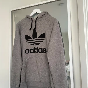 Grå Adidas hoodie med svart logga - Säljer en grå hoodie från Adidas med stor svart trefoil-logga och text på bröstet. Tröjan har huva med snörning och en klassisk känguruficka framtill. Perfekt för chill dagar eller när du vill ha en sportig look.