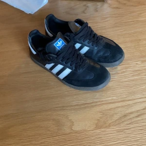 Adidas Samba svarta sneakers - Klassiska Adidas Samba sneakers i svart skinn och mocka med vita ränder och detaljer. Snygg retrostil med gummisula och svart snörning. Perfekta för dig som gillar streetwear och tidlös design. Blå Adidas-logga på plösen och guldfärgad Samba-text på sidan.