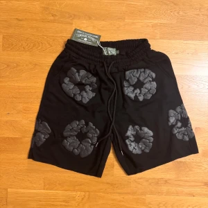 Svarta shorts från Denim Tears L - Svarta shorts från Denim Tears med unika gråa blommönster över hela plagget. De har elastisk midja med snörning och en bakficka. Sköna och stilrena, perfekta för dig som vill sticka ut med en cool streetwear-look.