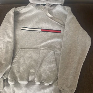 Grå hoodie Tommy Hilfiger - Snygg grå hoodie från Tommy Hilfiger med klassisk logga och ränder i rött, vitt och marinblått på bröstet. Tröjan har huva med dragsko och en stor magficka. Perfekt för en chill och stilren look.