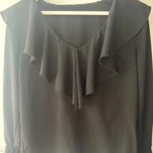 Svart blus med volang och rosetter - Svart blus med stor volang längs v-ringningen och knytrosetter vid ärmsluten. Lätt och flowigt tyg som ger en snygg siluett. Perfekt för dig som gillar detaljer och vill ha en stilren men ändå extra look.