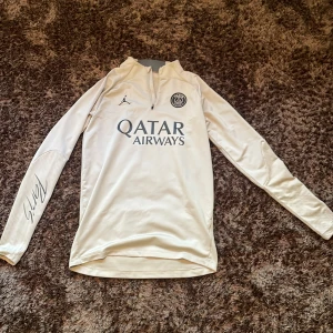 PSG långärmad träningströja  - Exklusiv PSG långärmad träningströja i ljusbeige med Jordan-logga och Paris Saint-Germain-märke på bröstet. Halv dragkedja framtill, tryckt Qatar Airways-sponsor och Ramos-signatur på ärmen. Slim fit-modell i mjukt, tekniskt material. Sitter som S-M.