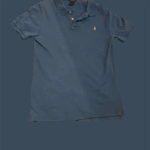 Blå pikétröja från Ralph Lauren - Klassisk blå pikétröja från Ralph Lauren med korta ärmar och gul broderad logga på bröstet. Tröjan har krage, två knappar och är gjord i mjuk bomull. Perfekt för en chill och stilren look.