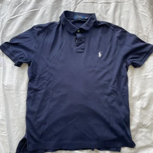 Marinblå pikétröja från Ralph Lauren - Säljer en klassisk marinblå pikétröja från Ralph Lauren i mjuk Pima-bomull. Tröjan har korta ärmar, krage och knappar framtill samt den ikoniska broderade loggan på bröstet. Perfekt för en clean och stilren look.