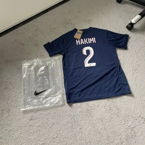 PSG Hakimi #2 Nike fotbollströja  - Snygg PSG fotbollströja med Hakimi #2 på ryggen. Tröjan är marinblå med röd och vit rand längs framsidan, PSG-logga och Nike Swoosh på bröstet samt Qatar Airways som sponsor. Tillverkad i lätt och ventilerande material, perfekt för match eller träning.