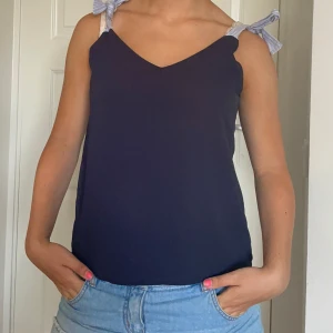 Mörkblå linnetopp med rosettband - Supersöt mörkblå linnetopp från Kristen Liz med v-ringning och axelband i blåvitrandigt tyg som knyts till rosetter. Perfekt till jeans eller kjol när du vill ha en chill men ändå snygg look. Luftig och lätt, passar grymt till varma dagar. Skriv om du undrar något 🩷