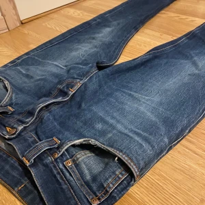 Blå jeans från Nudie Jeans Co - Säljer ett par klassiska blå jeans från Nudie Jeans Co med snygga slitningar och orangea kontrastsömmar. Jeansen har rak passform, fem fickor och är tillverkade i slitstarkt denimtyg. Perfekta för dig som gillar en tidlös och clean look. Modell: Steady Eddie. Står 33,34 inuti men sitter mer som 30,32