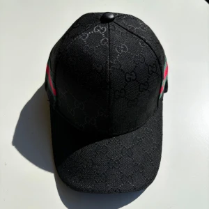 Svart keps från Gucci med GG-mönster - Snygg svart keps från Gucci med klassiskt GG-monogram över hela kepsen. På sidorna finns de ikoniska gröna och röda ränderna. Justerbar rem baktill och detaljerad insida med Gucci-logga. Perfekt accessoar för dig som gillar lyxig streetstyle.