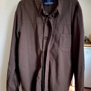 Human Scales light Jacket - Stilren brun skjorta från Human Scales i storlek M. Skjortan har klassisk krage, bröstficka och knäppning framtill. Tillverkad i mjuk bomull och passar perfekt till jeans eller chinos för en avslappnad look.