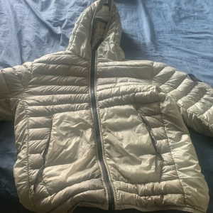 Jätte ljus grön dunjacka från Stone Island - Säljer en beige dunjacka från Stone Island med huva och klassisk patch på ärmen. Jackan har horisontella sömmar, två stora fickor framtill och dragkedja hela vägen. Perfekt för kalla dagar och riktigt snygg streetstil.