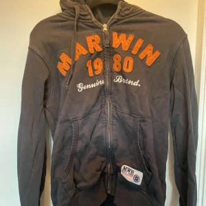 Svart hoodie från Marwin Sports M - Svart hoodie från Marwin Sports med orange broderad text 'MARWIN 1980' på bröstet. Tröjan har dragkedja, huva med snörning och två fickor framtill. Ribbstickade muddar och detaljer i orange och vitt på insidan av huvan. Perfekt för chill dagar.