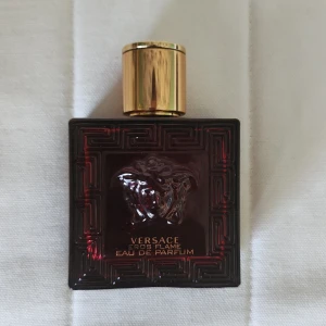 Versace Eros Flame Eau de Parfum -     Hälften kvar köptes på NK. Versace Eros Flame Eau de Parfum i en lyxig mörkröd glasflaska med guldigt lock. Flaskan har Versaces ikoniska Medusa-huvud i relief både på framsidan och på locket. Elegant och stilren design som verkligen sticker ut i parfymhyllan.