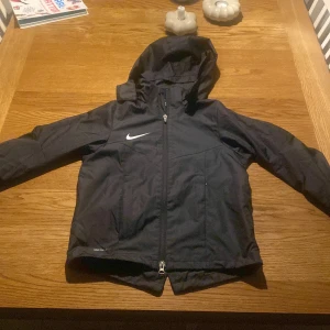 Svart Nike vindjacka med huva - Svart vindjacka från Nike med huva och dragkedja framtill. Jackan har Nike-logga på bröstet och är tillverkad i lätt polyester. Perfekt för blåsiga dagar och sportiga outfits. Enkel och stilren design som funkar till det mesta.