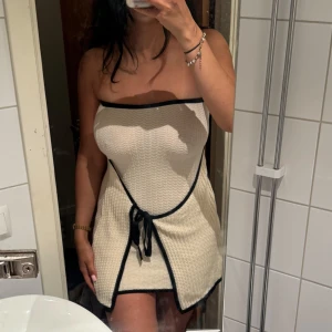 Stickad tubklänning med knytdetalj - Stickad kortklänning med svarta kanter och knytband framtill. Tubmodell utan axelband och med en omlottliknande detalj framtill. 