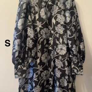 Svart blommig klänning med puffärm - Supersnygg svart klänning med stora grå blommor och puffiga långärmar. Klänningen har breda manschetter och ett elegant blommönster som verkligen sticker ut. Perfekt för dig som vill ha något unikt och trendigt!🌸