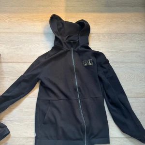 Svart EA7 hoodie med dragkedja - Svart hoodie från EA7 Emporio Armani med gul logga på bröstet och ryggen. Tröjan har hel dragkedja, huva och två fickor framtill. Tillverkad i mjuk bomullsmix, perfekt för en chill och stilren look.