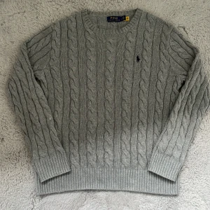 Grå kabelstickad tröja Polo Ralph Lauren  - Kabelstickad grå tröja från Polo Ralph Lauren med klassisk rund halsringning och broderad logga på bröstet. Tröjan är i mjuk stickad bomull och har ribbade muddar vid ärmslut och nederkant. Perfekt för lager-på-lager och chill stil. 20% rabbat under helgen 1100 kr❌ 880 kr✅