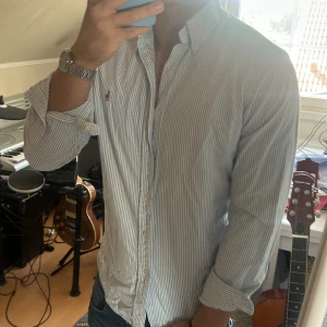 Randig skjorta från Ralph Lauren - Säljer en klassisk skjorta från Ralph Lauren med blå och vita ränder. Skjortan har lång ärm, knappar hela vägen och den ikoniska broderade loggan på bröstet. Tillverkad i bomull och har en snygg, normal passform. Perfekt för dig som gillar stilrena och tidlösa plagg.