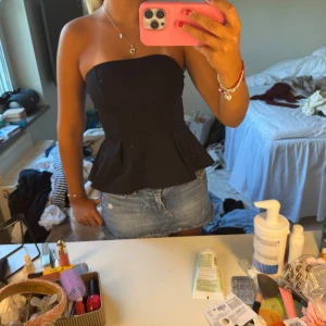 Svart peplum tubtopp - Snygg svart tubtopp med peplumvolang nertill. Toppen är axelbandslös och har en tight passform upptill som ger en stilren siluett. Perfekt att matcha med jeansshorts eller kjol för en trendig look.