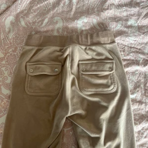 Beiga mjukisbyxor Juicy Couture XS - Går att pruta!!!Säljer ett par beiga mjukisbyxor från Juicy Couture i storlek XS. Byxorna har bred resår i midjan, två stora fickor med lock och knappar bak samt en sned ficka fram med broderad JC-logga. Mjukt material och relaxed fit.