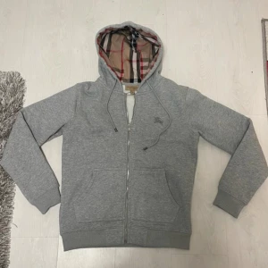 Grå Burberry hoodie  - Säljer en grå hoodie från Burberry med klassiskt rutigt foder i huvan och dragkedja framtill. 