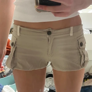 Beige shorts med fickor och snörning - Snygga beige shorts från Cargo Six med låga midja, stora sidfickor med knappar och cool snörning längst ner på benen. De är tillverkade i mjuk bomull och har vita detaljer vid midjan och bensluten. Perfekta för sommardagar när du vill ha en avslappnad och trendig look.