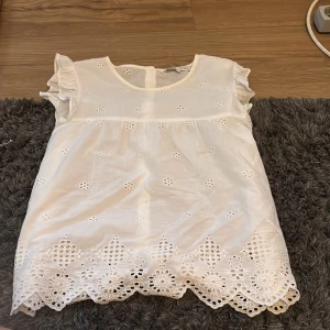 Vit blus med broderade detaljer - Supersöt vit blus med broderade blommor och volangärmar. Blusen har rund halsringning och vackert broderad nederkant med hålmönster. Perfekt för dig som gillar romantisk och luftig stil.
